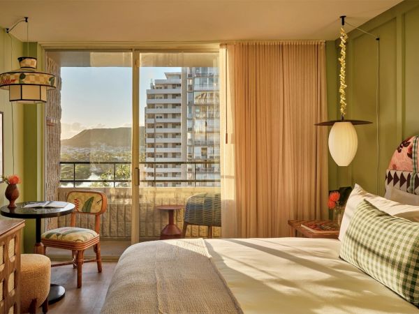 A cozy bedroom features a neatly made bed, a small dining table with chairs, and a large window with a balcony view of buildings and mountains.