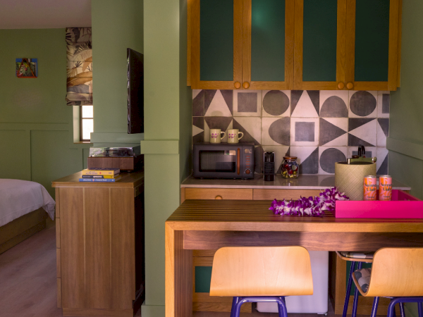 Dans une pi&egrave;ce peinte en vert, un coin cuisine compact avec un bureau, un &icirc;lot en bois, deux chaises claires, des &eacute;tag&egrave;res ouvertes, un micro-ondes et un plateau rose ; on aper&ccedil;oit en arri&egrave;re-plan une partie d'un lit et une petite fen&ecirc;tre.