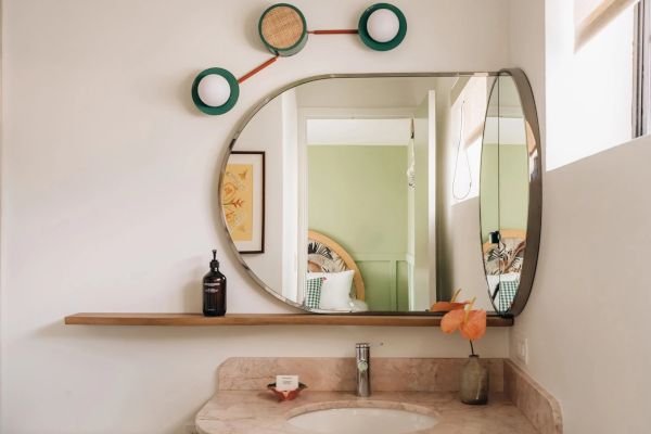 Bathroom with a unique mirror, shelf with a bottle, plant, soap, and wall decoration above the mirror. A bedroom is visible in the mirror.