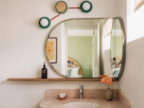 Bathroom with a unique mirror, shelf with a bottle, plant, soap, and wall decoration above the mirror. A bedroom is visible in the mirror.