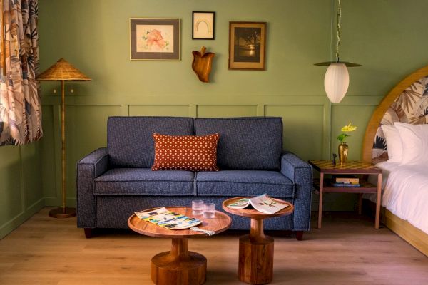 The image shows a cozy living space with a blue sofa, a small coffee table, framed pictures on the wall, a floor lamp, and a bed in the background.