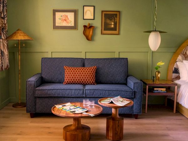 The image shows a cozy living space with a blue sofa, a small coffee table, framed pictures on the wall, a floor lamp, and a bed in the background.