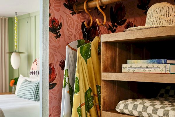 The image shows a cozy bedroom with a patterned pink wall, wooden hanger with clothes, shelves with a hat, books, and folded clothes.