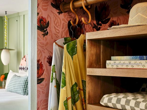 The image shows a cozy bedroom with a patterned pink wall, wooden hanger with clothes, shelves with a hat, books, and folded clothes.
