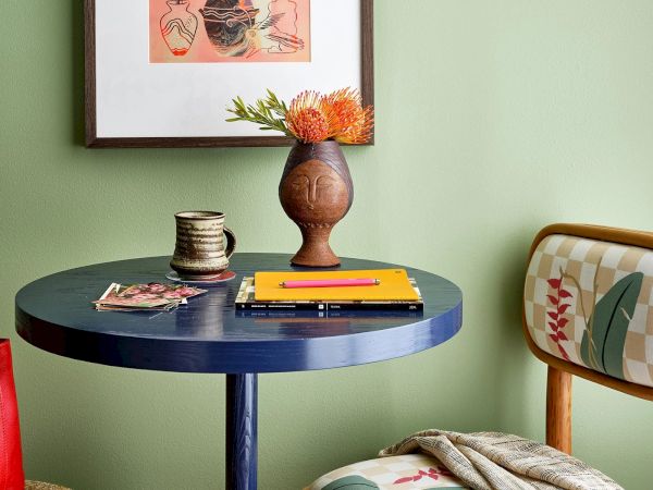 A small, round blue table with a vase of flowers, a notebook, and a mug. A red bag and a patterned chair are also visible in the scene.