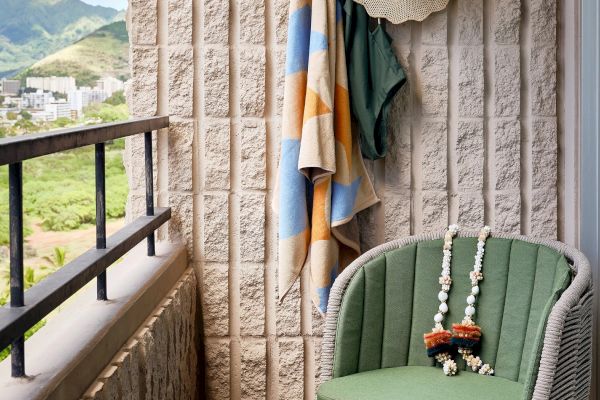 A cozy balcony with a green chair, colorful towels hanging, and a view of a city and mountains. A lei with flowers and beads is draped on the chair.