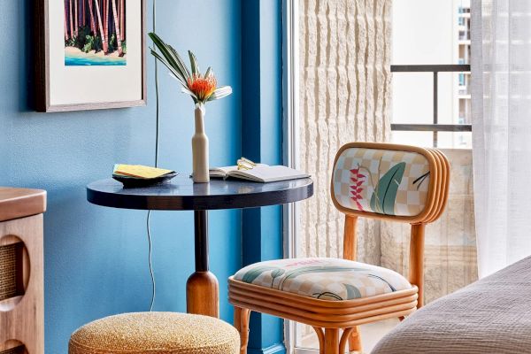 A cozy corner with a small round table, floral chair, ottoman, decorative vase with a flower, and an artwork on a blue wall.