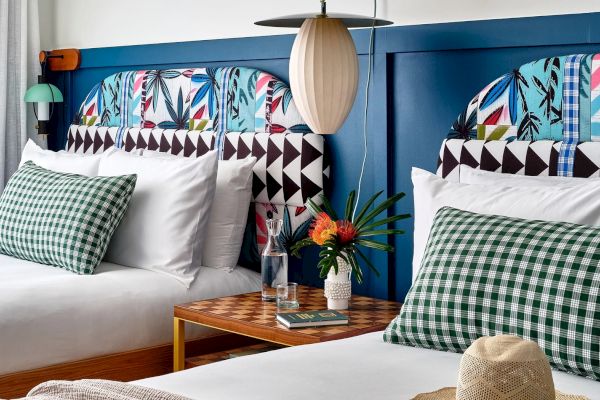 A hotel room features two double beds with colorful headboards, green checkered pillows, a side table with a lamp, flowers, and a hat.