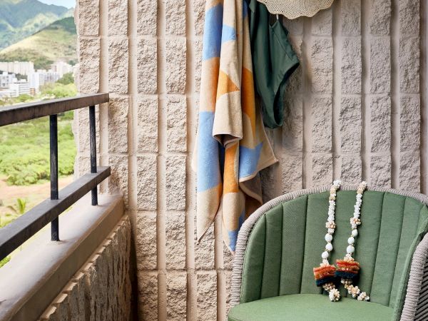 A small balcony with a green chair, colorful towels hanging, a hat, and two leis placed on the chair, overlooking a scenic landscape.