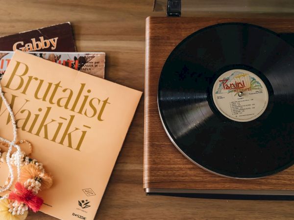 A vinyl record is on a wooden turntable, next to an album cover titled "Brutalist Waikiki" and other albums, with a colorful necklace on top.
