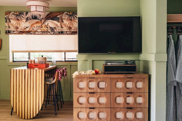 A cozy room features a patterned valance, wooden counter with stools, a modern dresser, and a wall-mounted TV, with aprons hanging on the side wall.