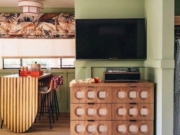 A cozy room features a patterned valance, wooden counter with stools, a modern dresser, and a wall-mounted TV, with aprons hanging on the side wall.
