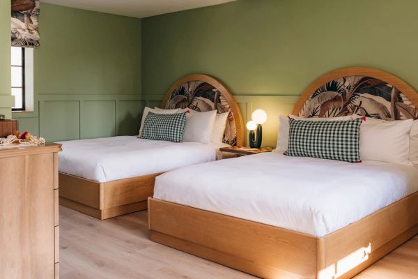 A cozy bedroom with two neatly made beds, wooden frames, checkered pillows, and a bedside table with a round lamp, all against green walls.