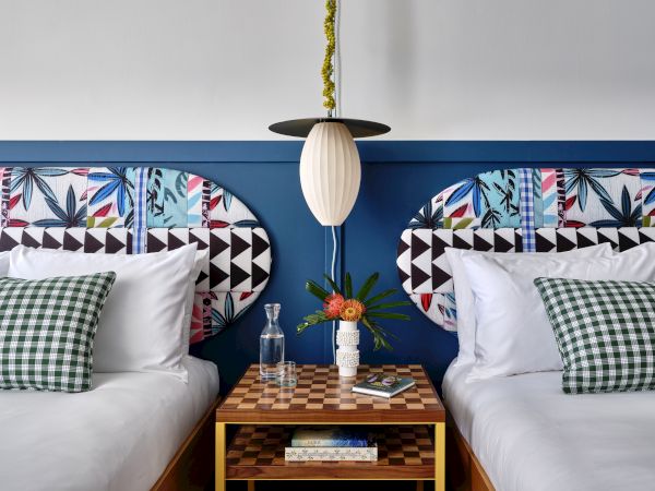 A bright bedroom with two beds, patterned pillows, a hanging lamp, and a small table with flowers and books.