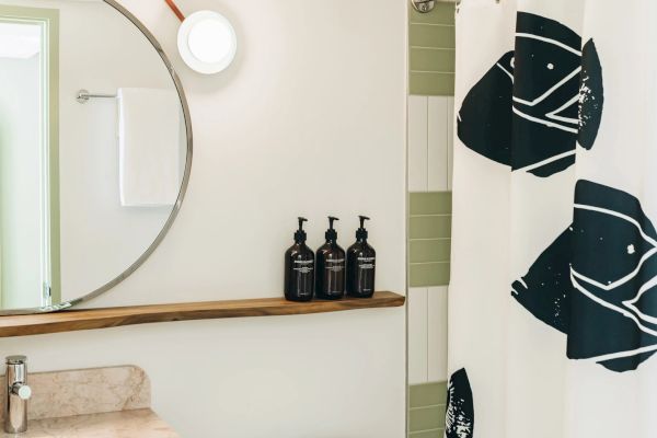 This image shows a bathroom with a round mirror, a beige sink, three black bottles on a wooden shelf, and a white shower curtain with black patterns.