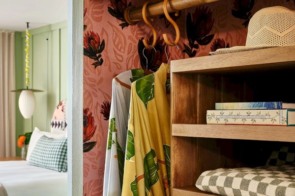 A cozy room with a bed, books on a shelf, a hat, and clothing hanging. The wallpaper features a floral pattern. The room has a warm and inviting vibe.