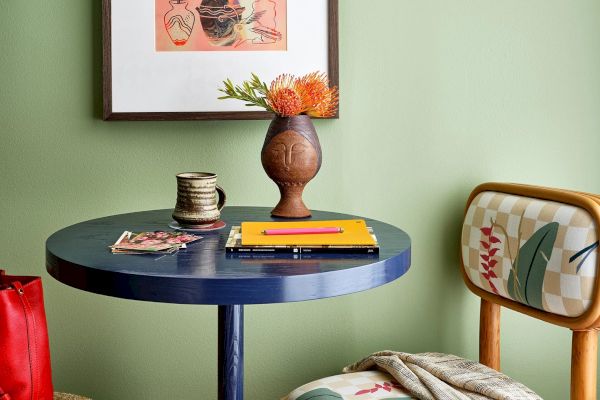 A small table with a notebook, pencil, vase with flower, mug, chair with cushions, and framed art on a green wall.