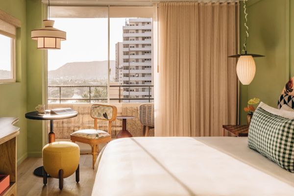 A cozy room with a bed, stylish lamps, a small table, chairs, and a balcony with a view of a distant mountain and nearby buildings, is seen.