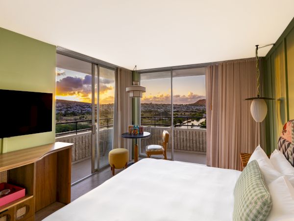Une chambre d'h&ocirc;tel moderne avec balcon, lit king-size, t&eacute;l&eacute;vision murale et vue sur la ville au coucher du soleil.