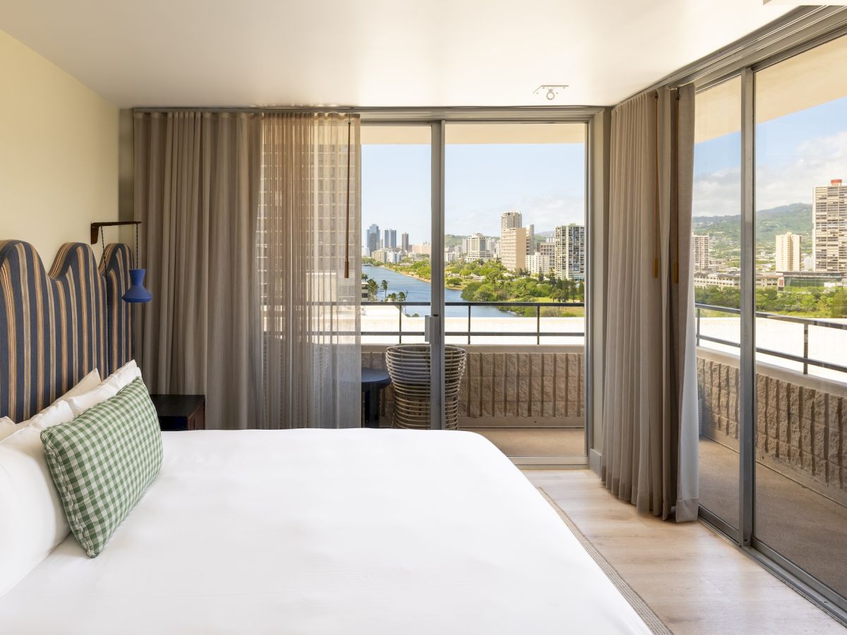 Una moderna habitaci&oacute;n de hotel con una cama grande, cortinas transparentes y una puerta corredera que deja ver el horizonte junto al r&iacute;o.