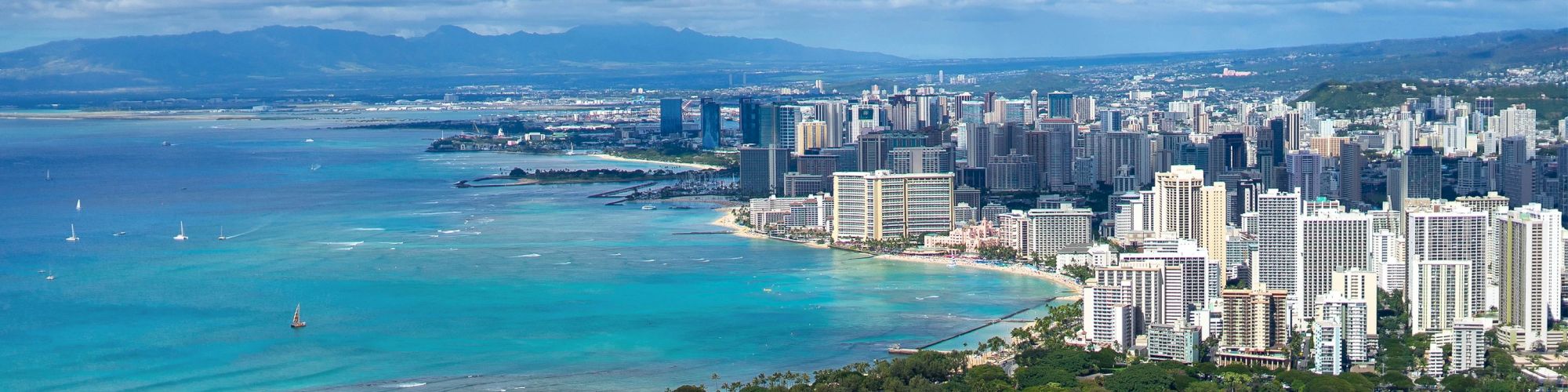 This image shows a coastal city with high-rise buildings beside a beautiful blue ocean, dotted with sailboats, and mountains in the background.