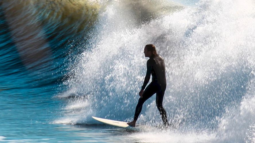 Un surfista con un traje de neopreno negro cabalga una ola, dejando una estela de agua blanca y salpicaduras; su silueta se recorta n&iacute;tidamente contra el oc&eacute;ano iluminado por el sol.