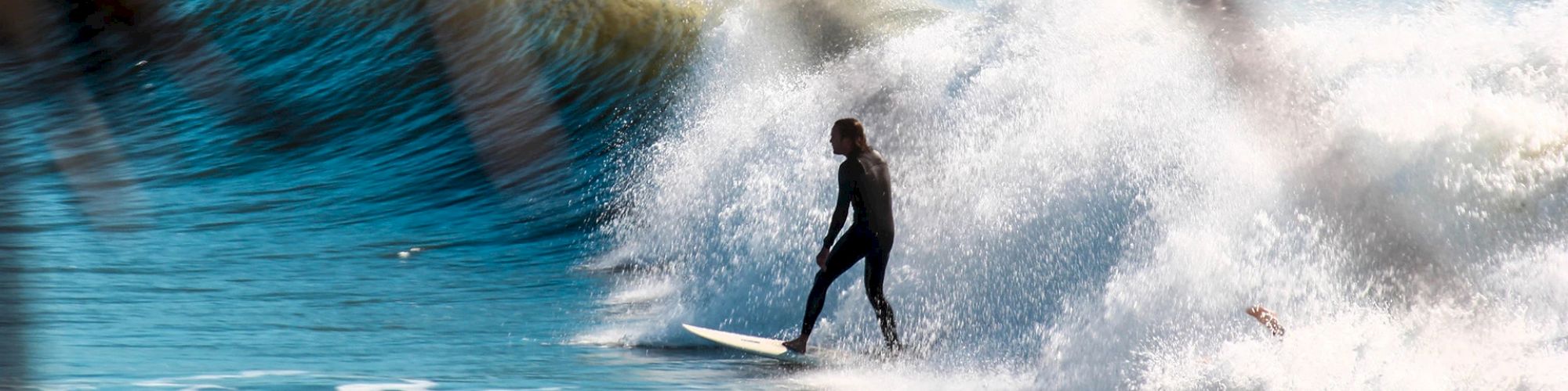 A surfer in a black wetsuit rides a wave, leaving a trail of white water and splashes; their silhouette is clear against the sunlit ocean.