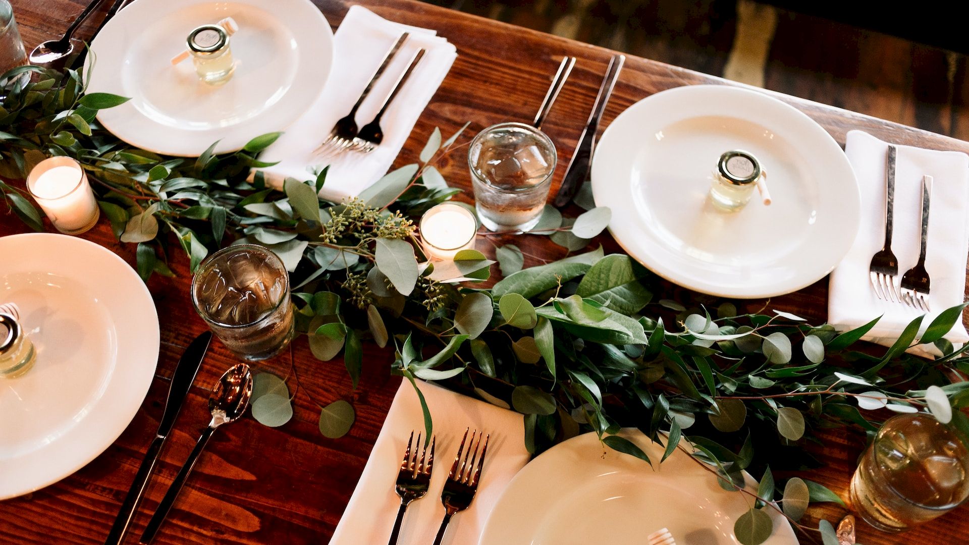 A table setting with white plates, silverware, glassware, candles, and a green leafy centerpiece on a wooden table.