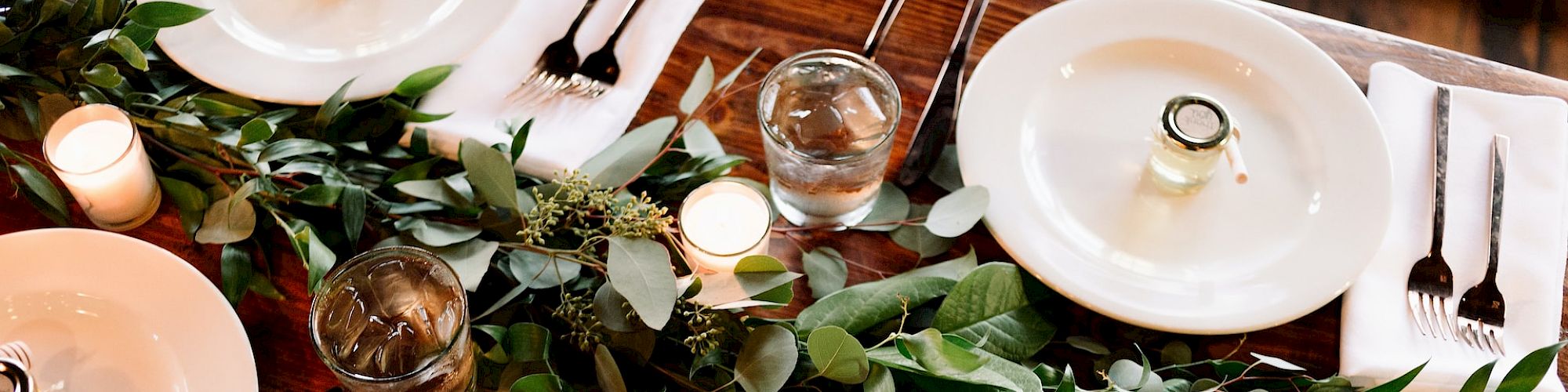 A table setting with white plates, silverware, glassware, candles, and a green leafy centerpiece on a wooden table.