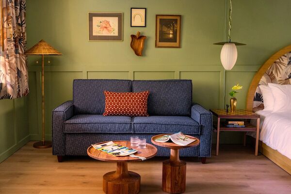 The image shows a cozy living space with a blue sofa, wooden coffee tables, wall art, a floor lamp, and a bed with a side table and a hanging lamp.