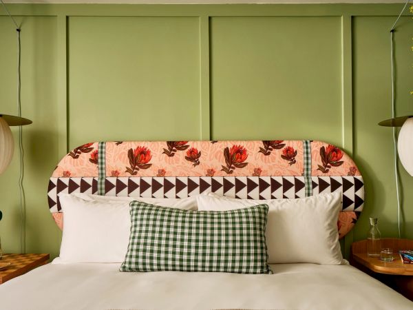 A bed with a patterned headboard, gingham pillow, and white sheets, flanked by two nightstands with decor, under pendant lights on green walls.