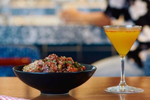 A bowl of food, possibly rice with toppings, is next to a yellow cocktail on a wooden table with a red-striped straw beside it.