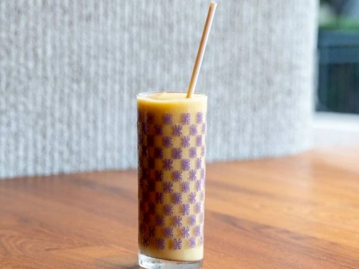 A tall, patterned glass filled with a yellow-orange beverage, topped with a straw, sits on a wooden surface.