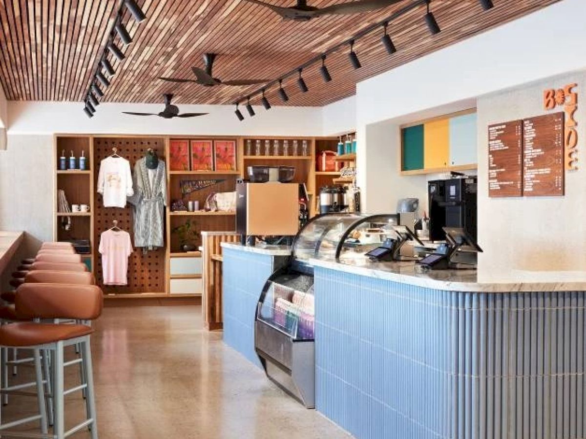 A modern café with a counter, stools, menu board, and merchandise shelving, featuring a minimalist design and ceiling fans.