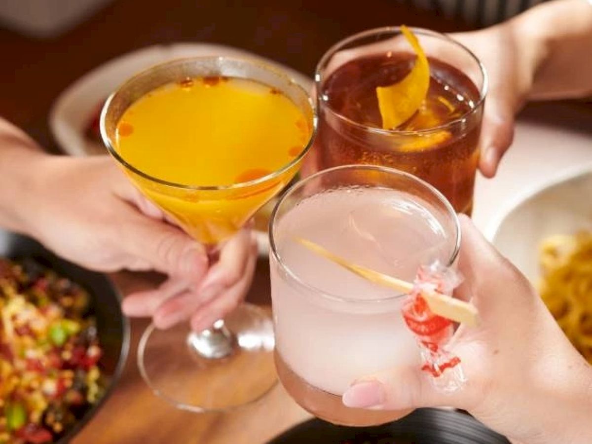 Three hands holding different cocktails are clinking glasses over a table with food dishes, suggesting a social gathering or celebration.