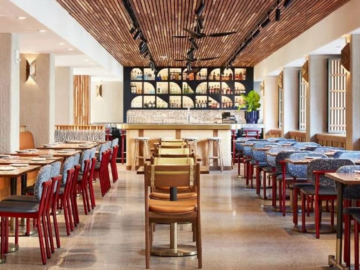 This image shows an empty, modern restaurant with wooden accents and a bar at the back, lined with wine bottles on a backlit shelf.