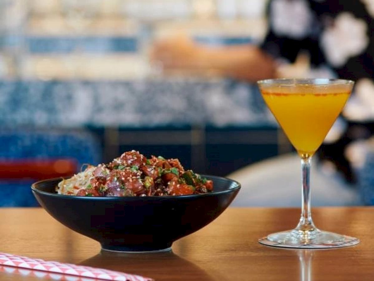 The image shows a bowl of food, perhaps a salad or poke bowl, and a yellow cocktail on a table with a striped straw next to it.