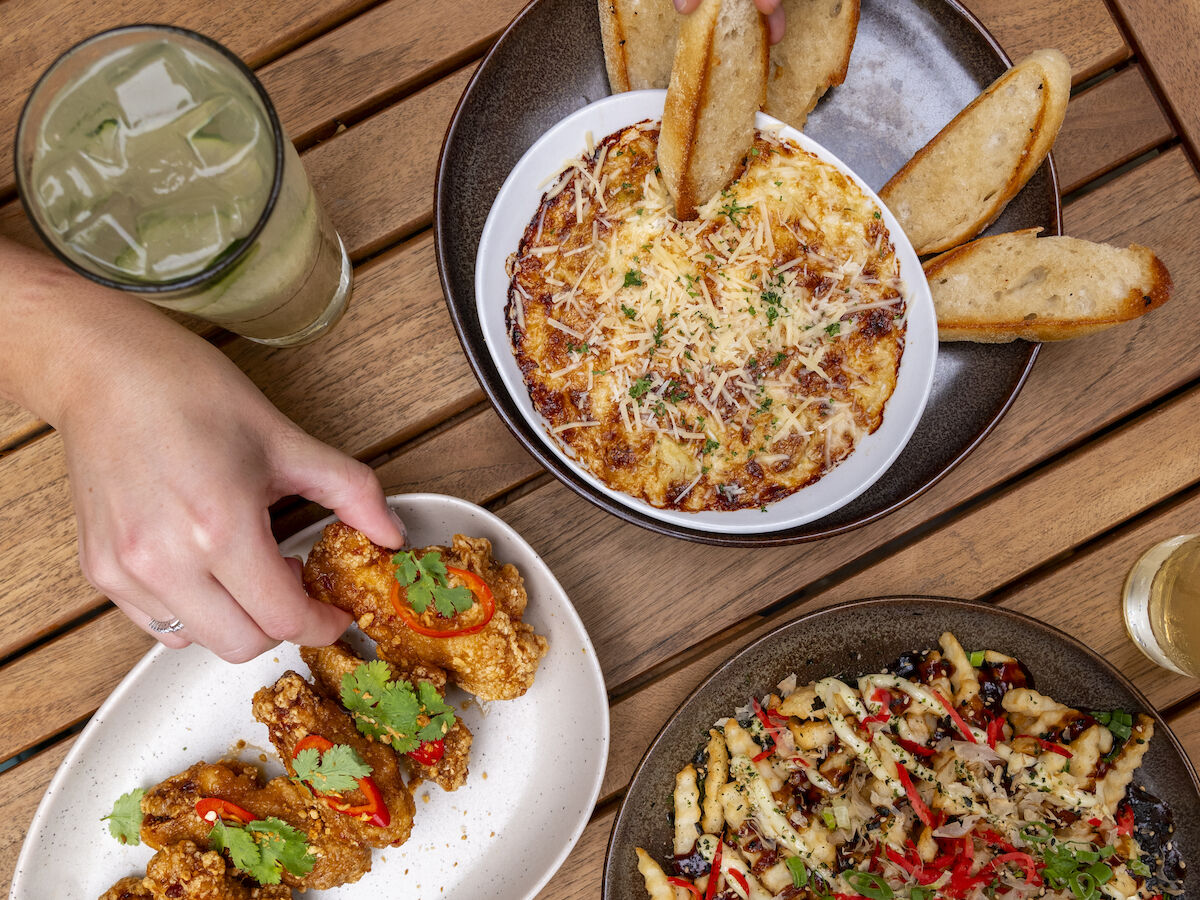 The image shows various dishes on a wooden table, including pasta, bread, chicken wings, and drinks. Two hands are seen reaching for the food.