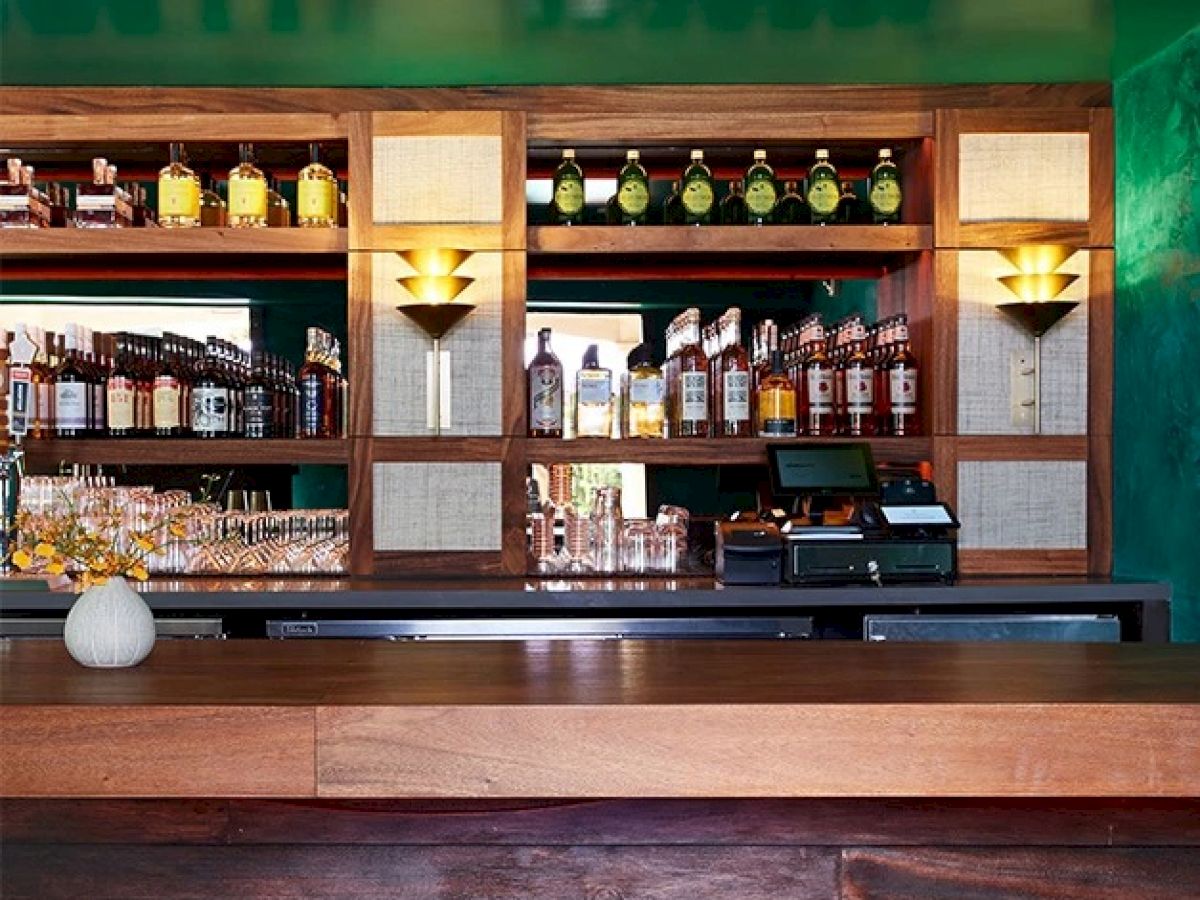 A bar counter with a wooden finish, backed by shelves stocked with various liquor bottles, wine glasses, and a cash register, in a warmly lit setting.