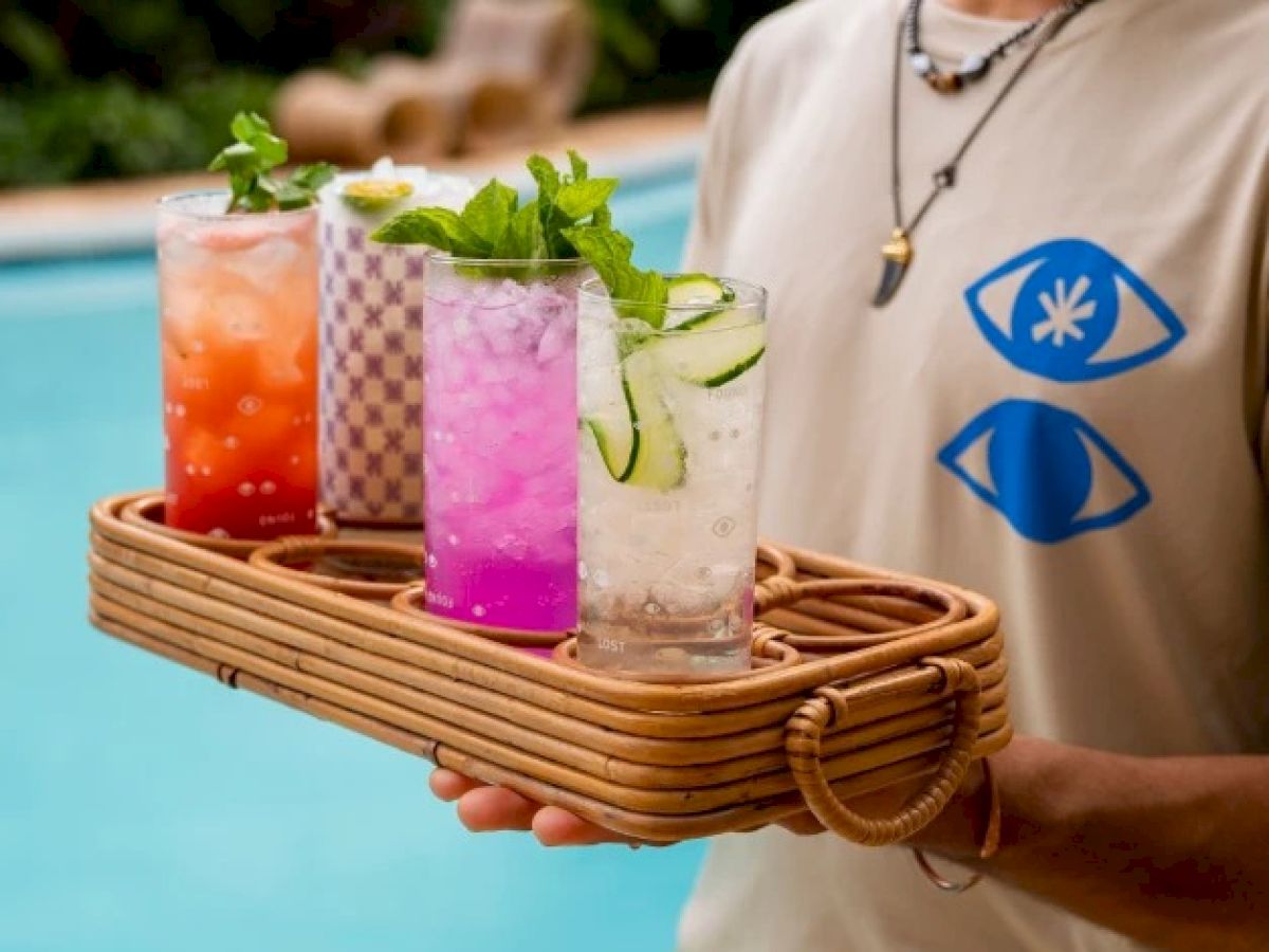 A person is holding a wicker tray with four colorful drinks, garnished with mint and cucumber, by a pool.