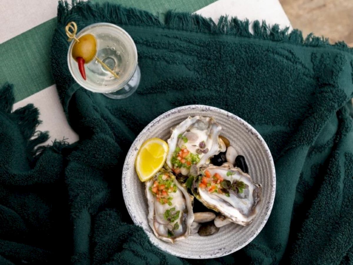 The image shows a bowl of oysters with a lemon wedge, garnished with herbs and vegetables, alongside a cocktail with an olive, on a green towel.