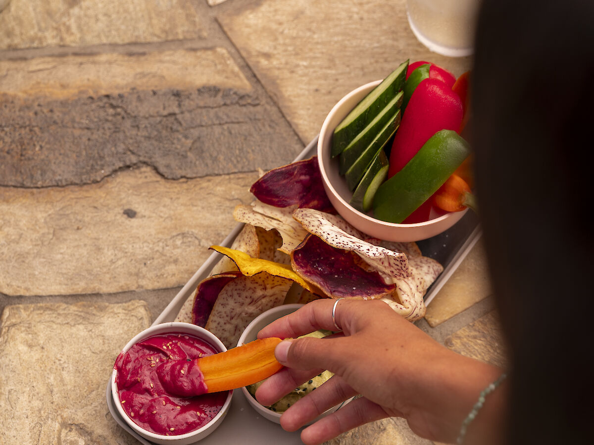 A hand reaching for food on a platter with sliced vegetables, meats, and two drinks placed on a stone surface outdoors, including colorful drinks.