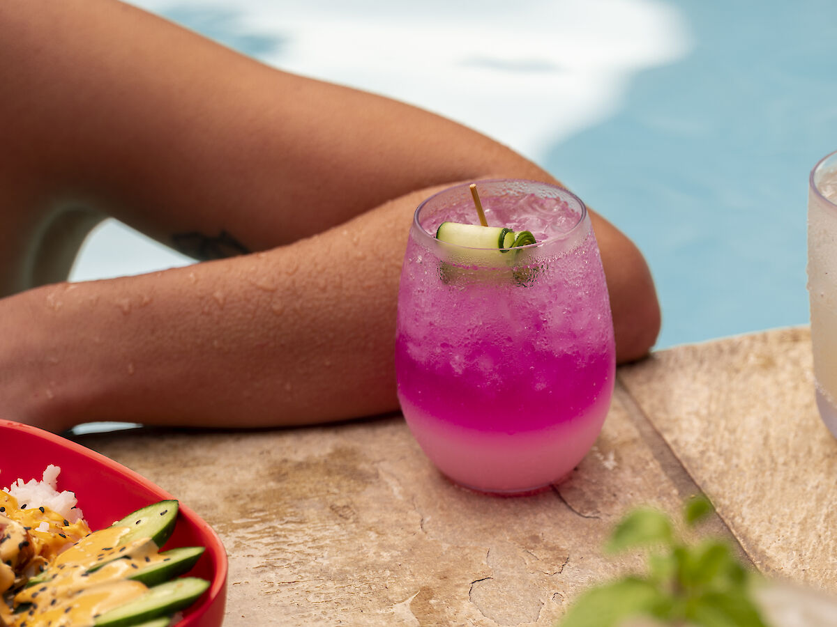 An arm rests by a poolside with vibrant drinks and food. There's a purple drink, a creamy drink garnished with mint, and sushi on a red platter.