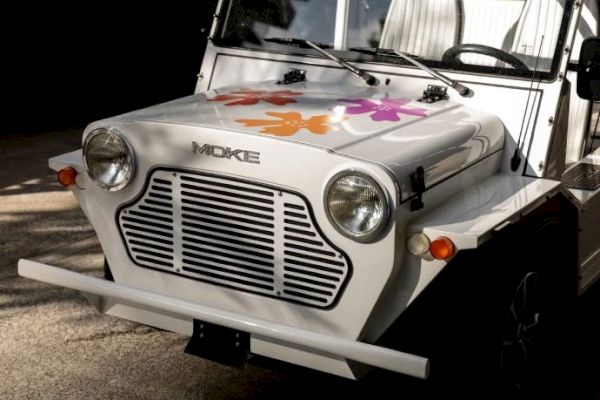 A white MOKE vehicle with a front floral decoration, headlights, and a distinctive grille, parked outdoors.