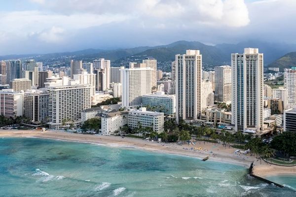 The image shows a coastal cityscape with tall modern buildings, a sandy beach, and turquoise ocean waters, set against a backdrop of mountains and cloudy skies.