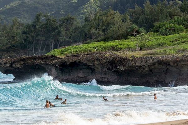 People are swimming and surfing in the sea near a rocky, lush green coastline with waves crashing onto the shore where sandy beach is visible.