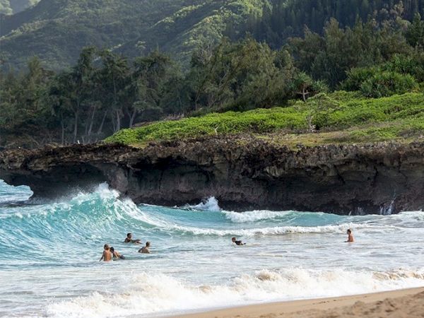 People are swimming and surfing in the sea near a rocky, lush green coastline with waves crashing onto the shore where sandy beach is visible.