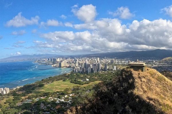 A scenic view of a coastal city with a large grassy area, buildings, and shoreline under a partly cloudy sky, with a structure on a hill.