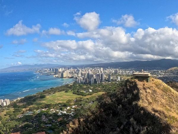 A scenic view of a coastal city with a large grassy area, buildings, and shoreline under a partly cloudy sky, with a structure on a hill.