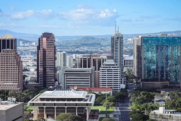 A cityscape featuring tall buildings, including modern skyscrapers, with mountains and a partly cloudy sky in the background.